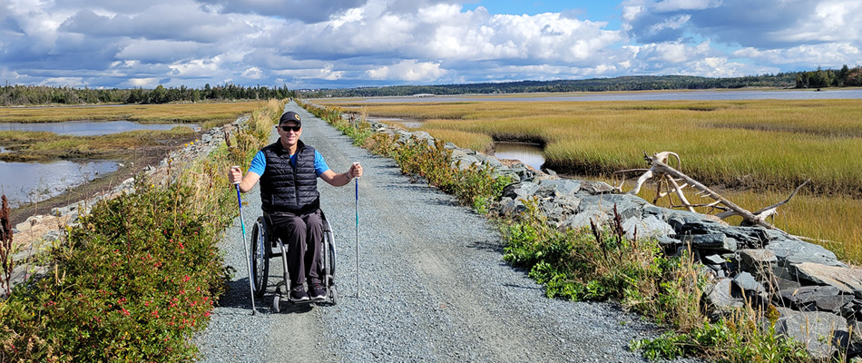 Un homme en fauteuil roulant sur un sentier en gravier compacté avec un bâton de randonnée dans chaque main. De chaque côté, il y a des marais. 