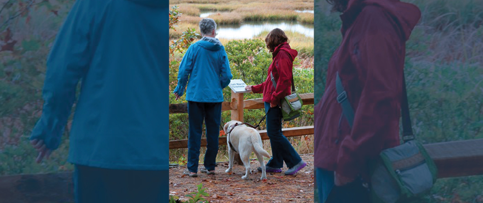 Deux adultes, dont l'un tient un chien en laisse, devant un panneau qui comprend du braille.