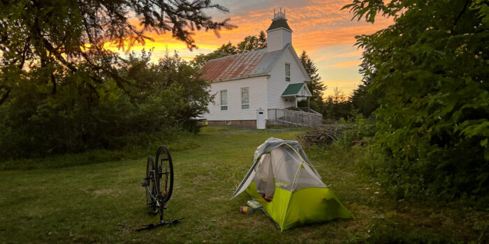 A tent and a bike on the grass in front of an older house with a sunset in the background
