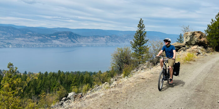 A man biking along a dirt trail on a mountain ridge with expansive views of a large lake and densely forested hills in the background