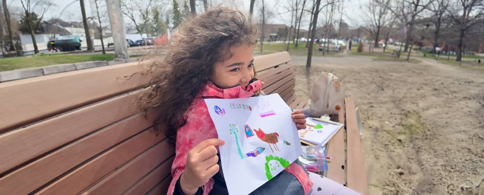 Selena holding her drawing up while sitting on a park bench | Selena tient son dessin alors qu'elle est assise sur un banc de parc