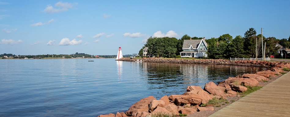 Boardwalk along water with lighthouse in distance at Charlottetown beach. | Une promenade pittoresque le long de la plage de Charlottetown, menant à un phare lointain.