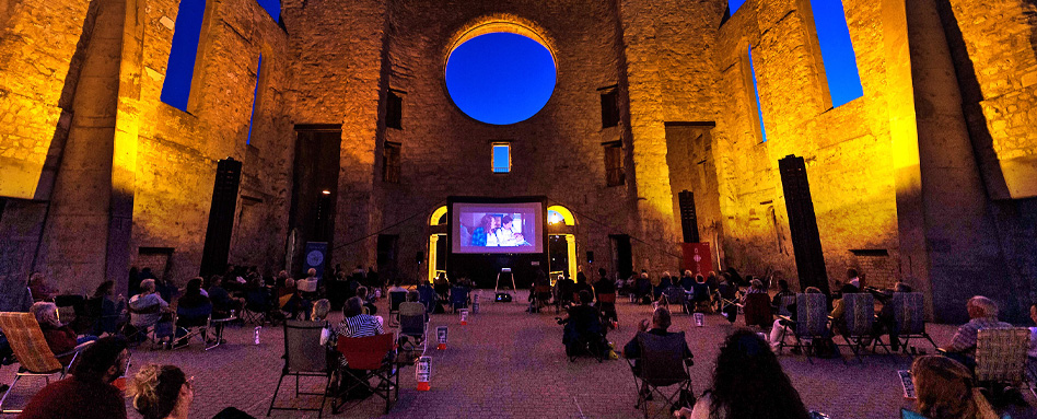 Group of people sitting in chairs in a courtyard at night, enjoying a movie at Cinéma sous les étoiles. | Groupe de personnes assises sur des chaises dans une cour la nuit, regardant un film au Cinéma sous les étoiles. 