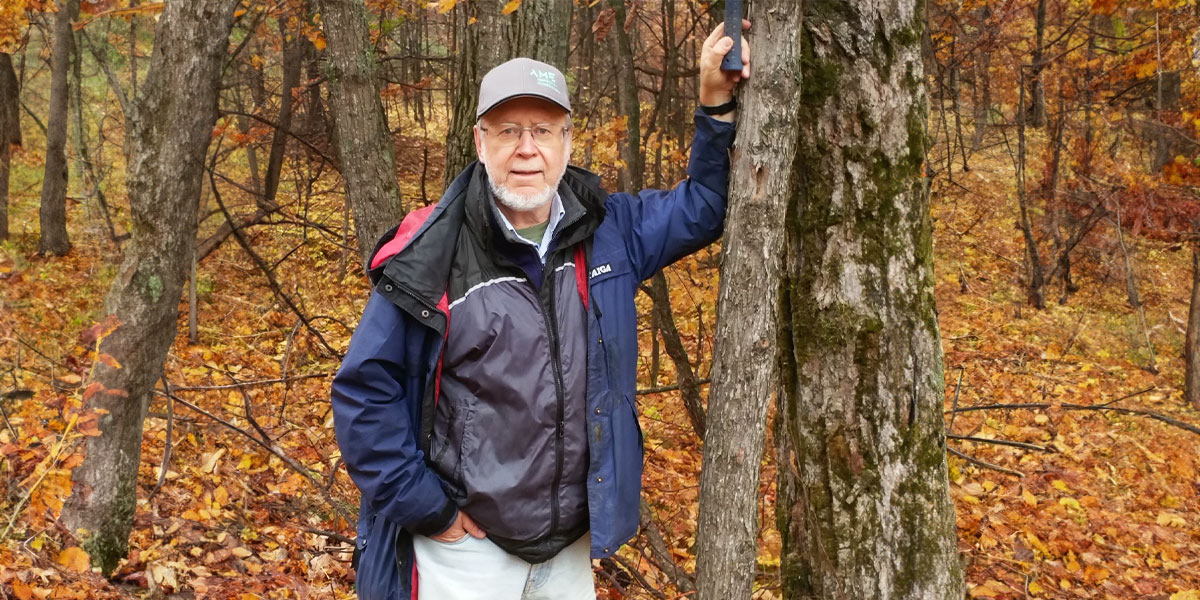 Edward Balon, stands beside a tree in a serene wooded area, surrounded by nature's tranquility. Edward Balon, stands beside a tree in a serene wooded area, surrounded by nature's tranquility.