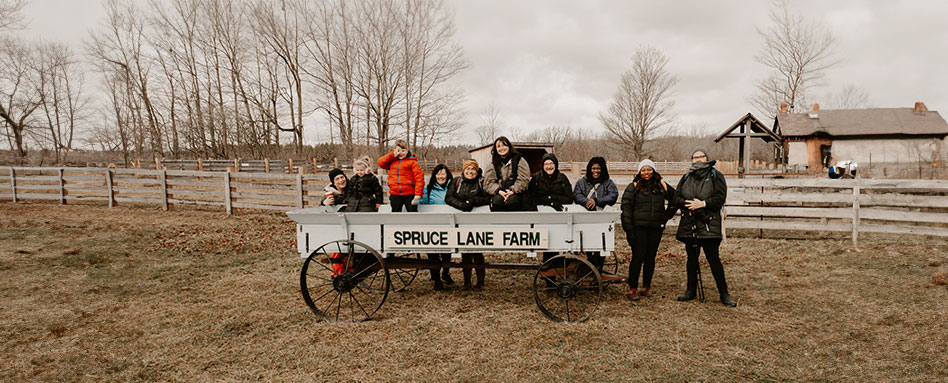 A diverse group of individuals stands together in front of a wagon at Spruce Lane Farm. Un groupe diversifié d'individus se rassemble devant un chariot à la ferme Spruce Lane. 