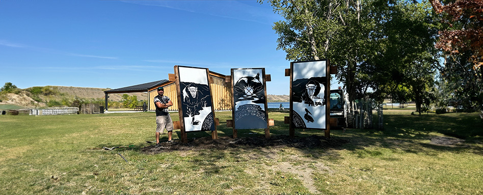 Harley Sinclair, a Peepeekisis First Nation artist, stands next to three large black and white posters displayed on a grassy field, highlighting Indigenous culture in Echo Valley Provincial Park. Harley Sinclair, une artiste de la Première Nation Peepeekisis, se tient à côté de trois grandes affiches en noir et blanc exposées sur un terrain gazonné, mettant en valeur la culture autochtone du parc provincial Echo Valley.