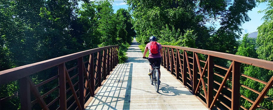 A cyclist traverses a bridge over a lush forest along the Goderich Rail Trail, enjoying the scenic view. Un cycliste traverse un pont au-dessus d'une forêt luxuriante le long du Goderich Rail Trail, profitant de la vue panoramique.