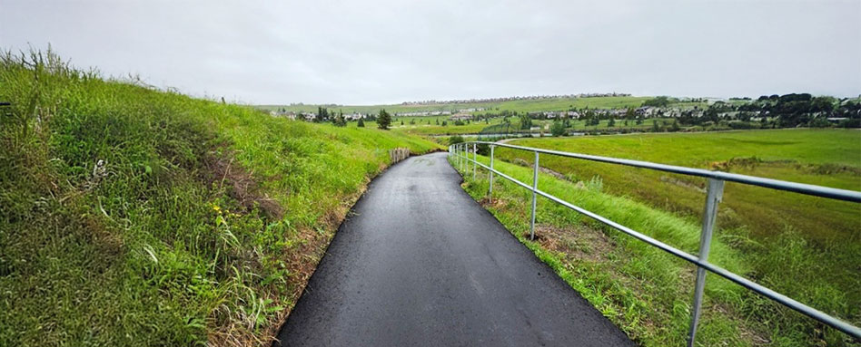 A paved path winds through the Town of Cochrane Trail, leading to a grassy hillside adorned with trees. Un sentier pavé serpente à travers le sentier de la ville de Cochrane, menant à une colline herbeuse ornée d'arbres.