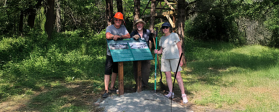 Three individuals stand beside a tree, featuring wayfinding signage in Haldimand County. Trois personnes se tiennent à côté d'un arbre, avec une signalisation d'orientation dans le comté de Haldimand.