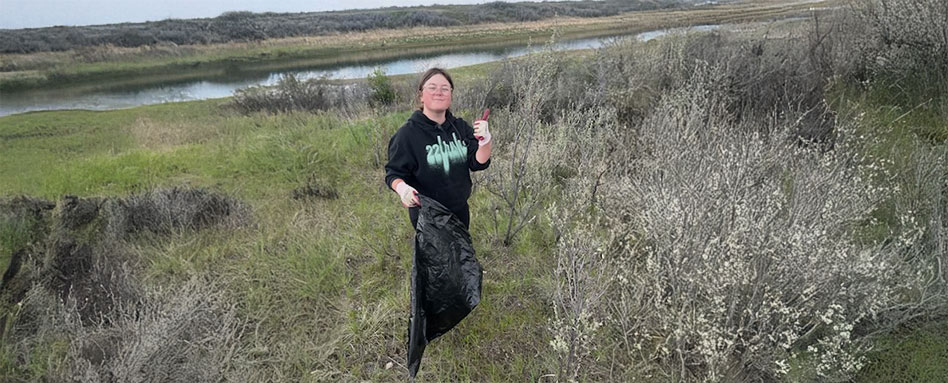 Taylor stands in a field, holding a black garbage bag. Taylor se tient dans un champ, tenant un sac poubelle noir. 