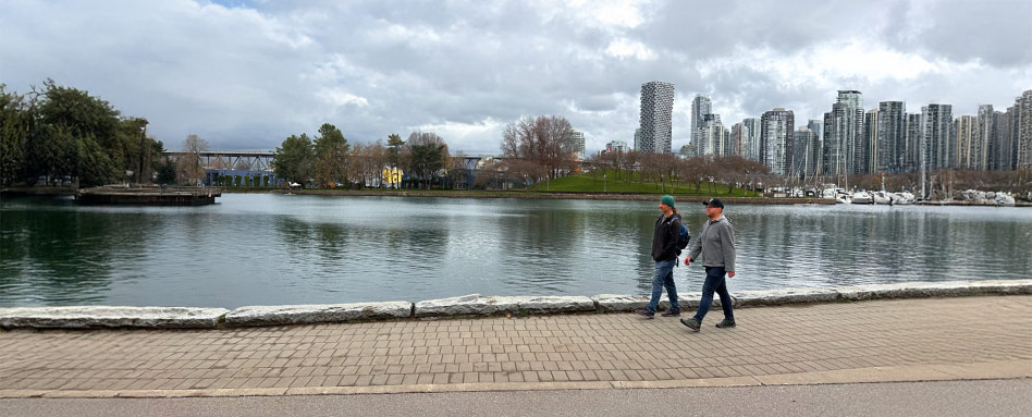 Two individuals walking by a lake with a city skyline in the background, near Stanley Park Seawall, Vancouver. Deux personnes marchent au bord d’un lac, avec la silhouette urbaine de la ville en arrière-plan, près du mur de soutènement du parc Stanley, à Vancouver.