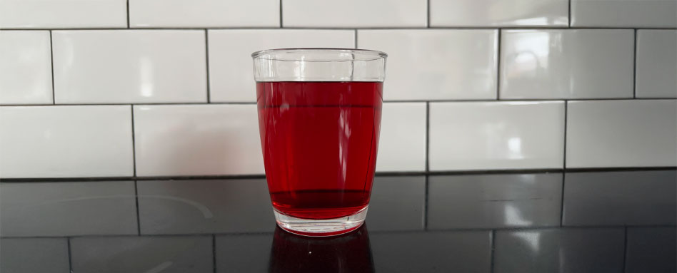 A glass of vibrant hibiscus tea resting on a countertop, showcasing its rich red hue. Un verre de thé à l’hibiscus d’une teinte rouge vive, posé sur un comptoir.