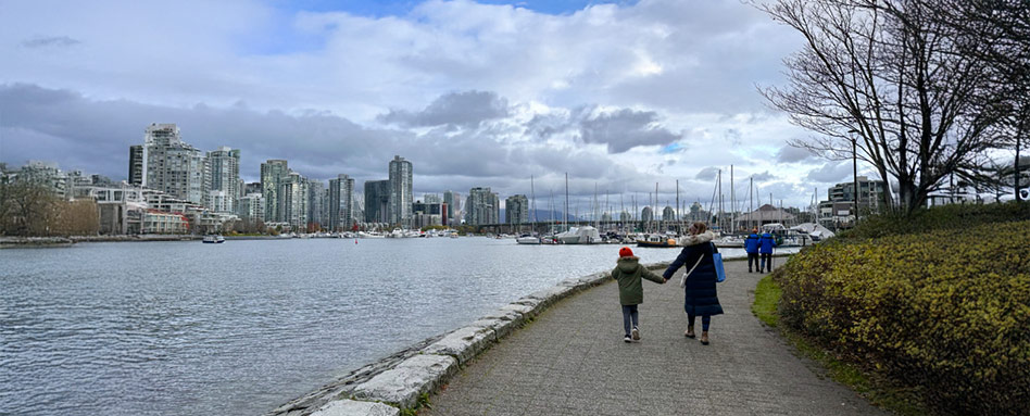 Two individuals stroll along the False Creek Seawall path with the Vancouver skyline and buildings visible in the background. Deux personnes se promènent le long du sentier du mur de soutènement de False Creek, avec la silhouette urbaine de Vancouver et ses bâtiments en arrière-plan.