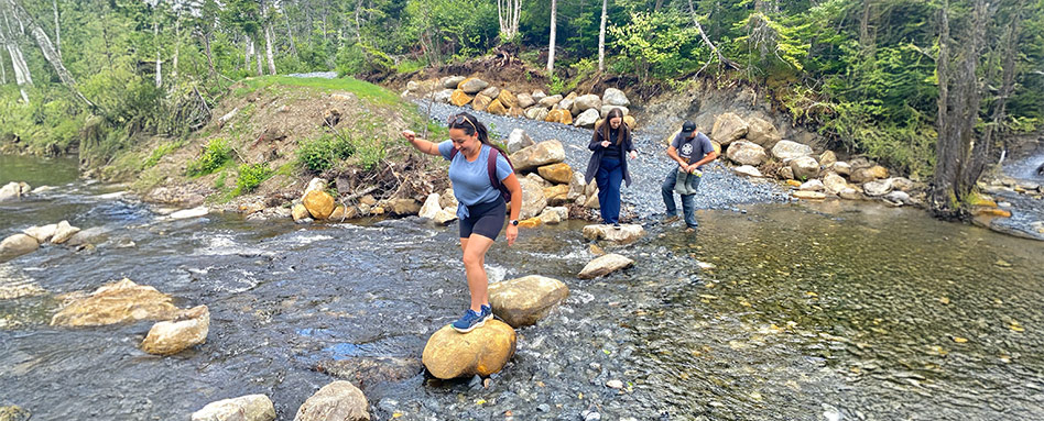 Three people navigate a rocky stream at the Ophiolite Massif Geological Site, highlighting the geological features of the area. Trois personnes traversent un ruisseau rocheux sur le site géologique du Massif ophiolitique, mettant en valeur les caractéristiques géologiques de la région.