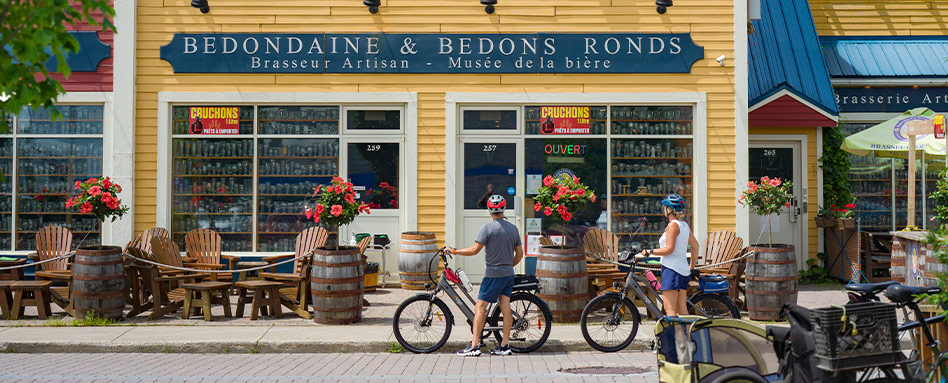 In Quebec's Montéregie region, a man and woman cycle past a store, showcasing a vibrant outdoor lifestyle. Dans la région de la Montérégie, au Québec, un homme et une femme à vélo passent devant un commerce, illustrant un mode de vie dynamique en plein air.