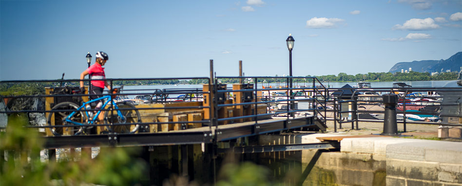 A woman riding a bicycle on a bridge over the Chambly Canal, surrounded by scenic views. Une femme à vélo traversant un pont au-dessus du canal de Chambly, entourée de paysages pittoresques.