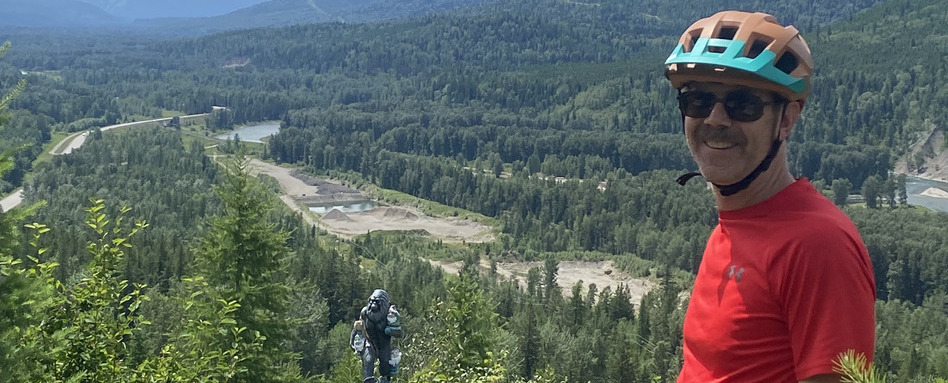man smiling at camera wearing a bike helpmet overlooking a tree-filled valley