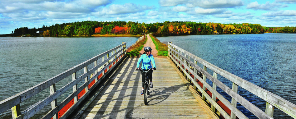 Cyclist riding along a wooden bridge over blue waters on the Confederation Trail in Prince Edward Island | Personne à vélo traversant un pont de bois au-dessus d’eaux bleuâtres sur le Sentier de la Confédération, à l’Île-du-Prince-Édouard.