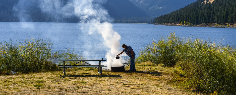 man putting out a bonfire with lake and mountains behind