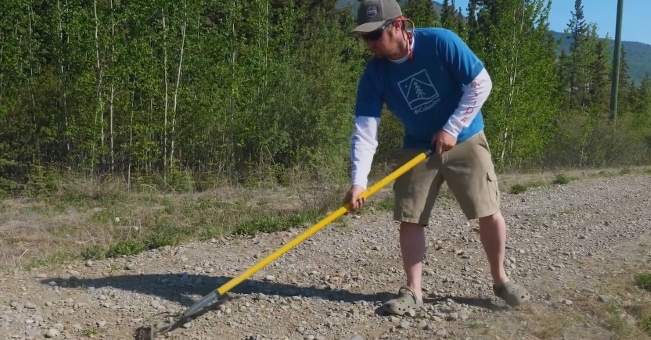 A person rakes gravel on a trail with trees in the background.