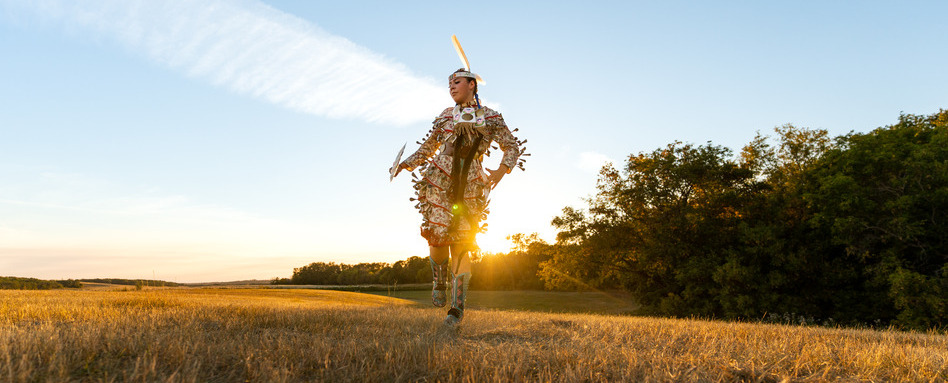 indigenous person in tradtional outfit dancing in field at sunset