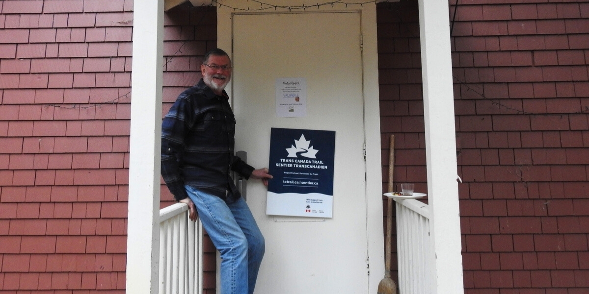 00Feature image_Volunteer recognition Slocan Valley Rail Trail | Sentier Transcanadien man standing infront of a door pointing at a sign and smiling