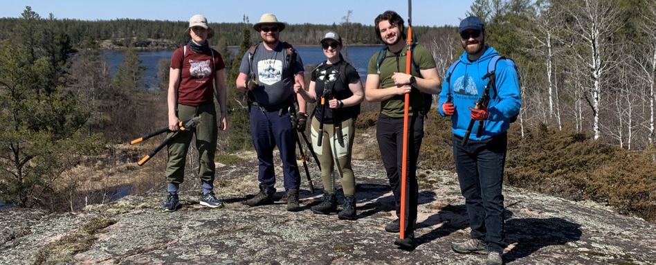 group of people in hiking and planting gear standing on hill on sunny day with water and forest behind