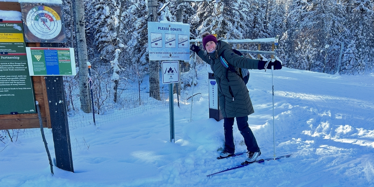 00Feature image_Emilie Brillon_AB_West Bragg Creek | Sentier Transcanadien woman smiling on skis in the snow pointing to a sign