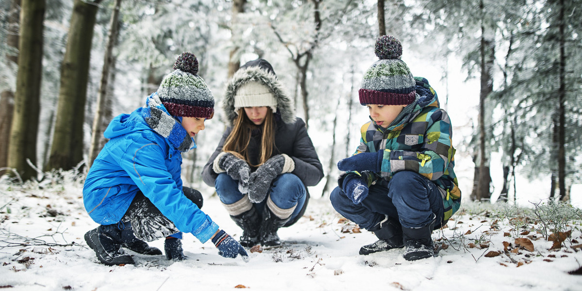 Three children crouching down and looking at something on the ground. The ground is covered in snow and the children are wearing blue winter suits.