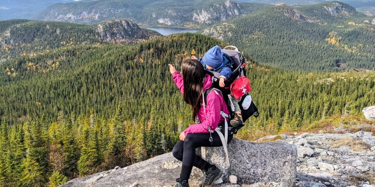 00Feature image_QC_Web story_Famille en Charlevoix_credit Naomie Veillette - ActivMom | Sentier Transcanadien A person sitting on a rock with a baby in a backpack on their back. They are looking at the mountain and valley view of evergreen trees.
