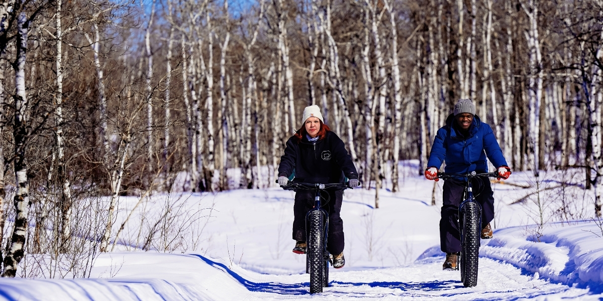 00SK_Pemiska_Feature image_fat bike_Credit Pemiska Tourism | Sentier Transcanadien Two people looking forward, riding fat-tire bikes along a trail in the winter
