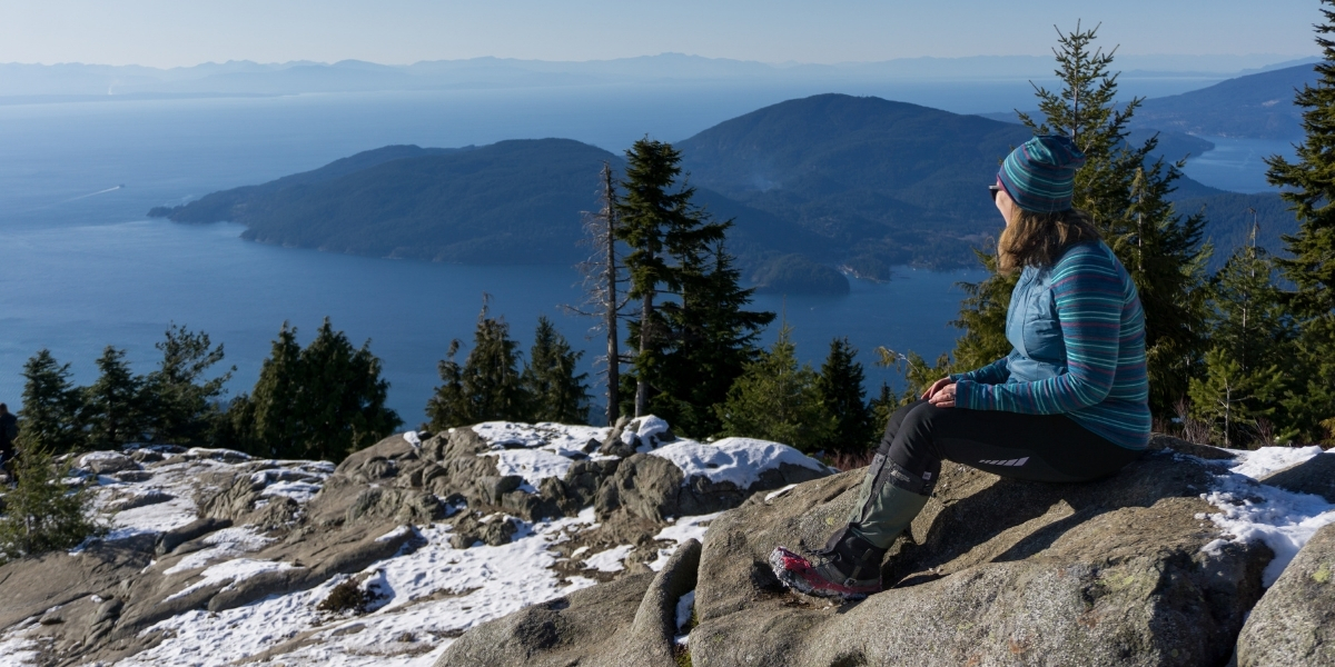 woman sitting atop a snowy montain on a sunyn day overlooking lake and mountains