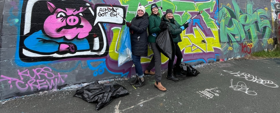 three women smiling with trash bags infront of graffiti wall