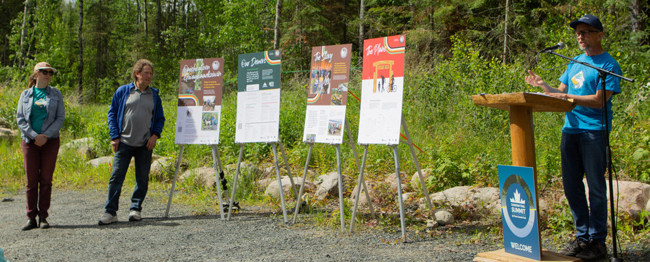 speakers stand at a podium infront of signage and forested area