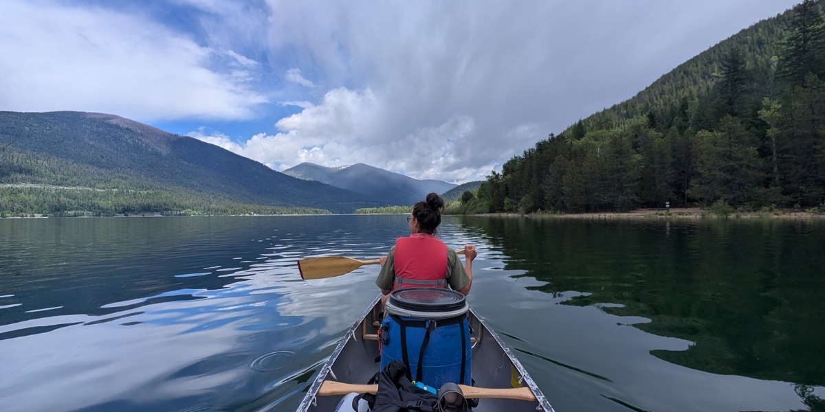woman in a canoe paddling on water between mountains and forest