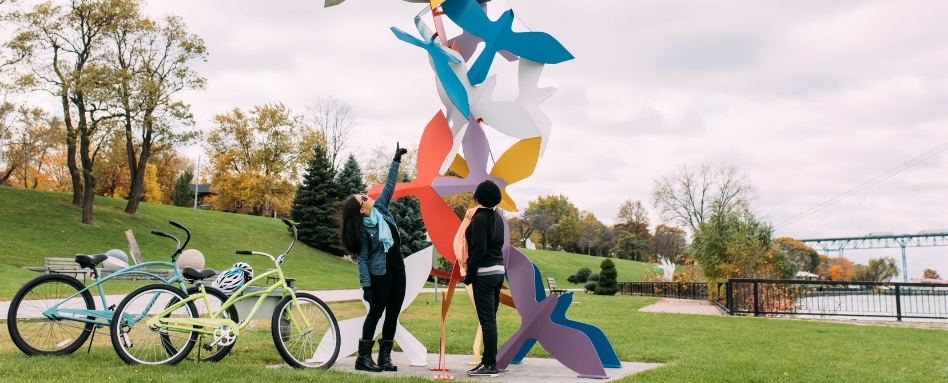 02Support image_ON_TWEPI article_Windsor sculpture garden_credit Tourism Windsor Essex Pelee Island | Sentier Transcanadien people standing infront of a statue waterfront with bikes and bridge behind
