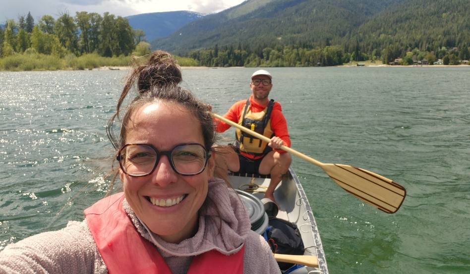 two people taking a selfie in a canoe on water with mountains behind