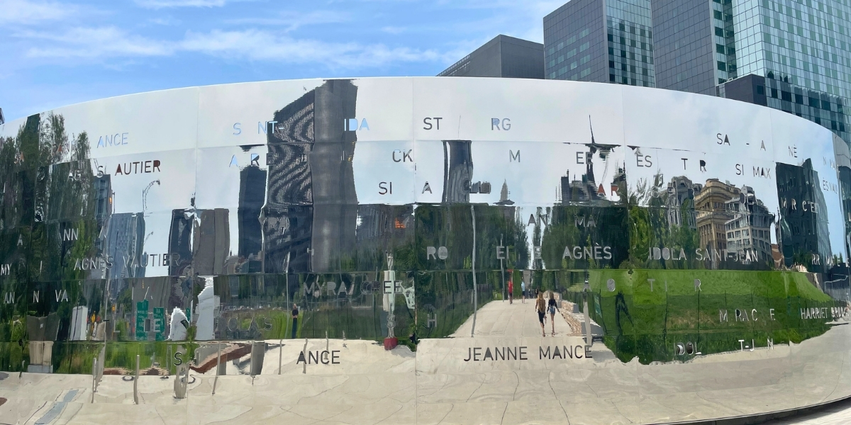 Curved mirrored metal wall reflecting buildings and visitors, with names etched across its surface in a public plaza: Jeanne Mance visible in letters.