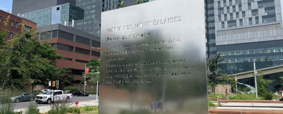 Reflective stainless steel memorial panel with French inscription 'Devoir de mémoire' in a city plaza with trees and modern office buildings nearby.